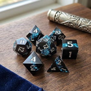 Op de afbeelding: Set van zeven polyhedrale dobbelstenen in zwart met blauwe accenten en zilveren cijfers. De dobbelstenen liggen op een houten oppervlak, met een decoratief metalen object op de achtergrond. Ideaal voor tabletop games.
