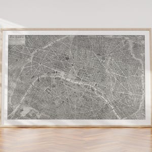 Peut inclure: Une carte en noir et blanc encadrée de Paris, France, exposée sur un sol en bois contre un mur blanc. La carte, dans un cadre en bois clair, montre la disposition complexe des rues de la ville. Le titre de la carte est visible dans le coin supérieur gauche.
