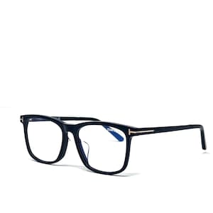 Peut inclure: Lunettes rectangulaires noires avec des verres transparents. Les montures présentent un design subtil et moderne avec un fin accent doré sur les branches. Les lunettes sont posées sur un fond blanc.