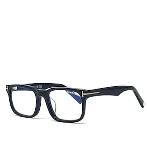 May include: Black rectangular eyeglasses with a gold accent on the temples. The glasses have a thick frame and clear lenses. The Tom Ford logo is visible on the left temple.