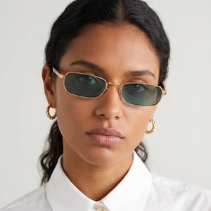 Peut inclure: Lunettes de soleil à monture dorée avec verres rectangulaires. Les lunettes ont une fine monture dorée et des verres vert foncé. Le modèle porte des créoles dorées et une chemise blanche. L'image est un gros plan.