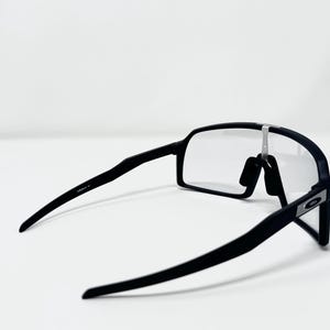 May include: Black sports sunglasses with clear lenses. The sunglasses have a sleek, modern design with a matte finish. The frame is black, and the lenses are rectangular. The arms of the sunglasses are thin and curve slightly.