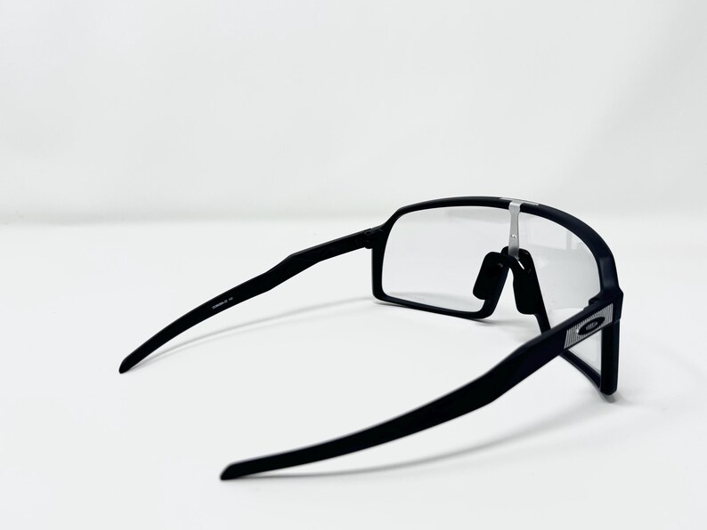 May include: Black sports sunglasses with clear lenses. The sunglasses have a sleek, modern design with a matte finish. The frame is black, and the lenses are rectangular. The arms of the sunglasses are thin and curve slightly.