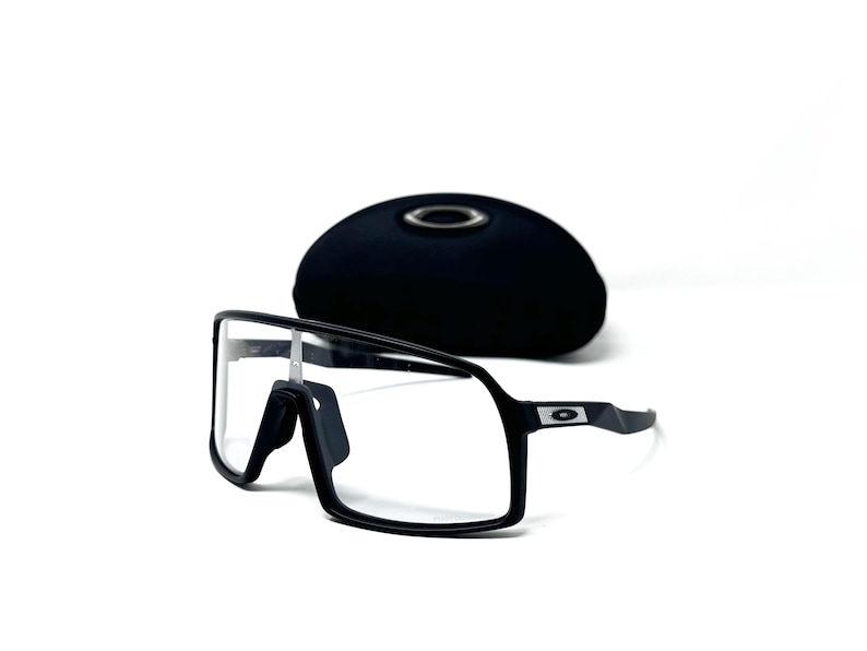 May include: Black framed sunglasses with clear lenses and a black case. The sunglasses have a modern, sporty design with a rectangular shape. The case is oval and has a silver emblem.