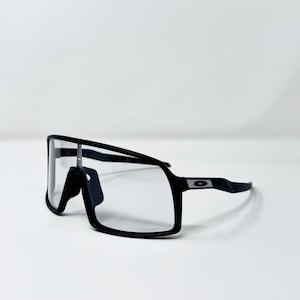 May include: Black sports sunglasses with a rectangular frame and clear lenses. The sunglasses have a sleek, modern design with a black frame and a small logo on the temple. The glasses are set against a white background.