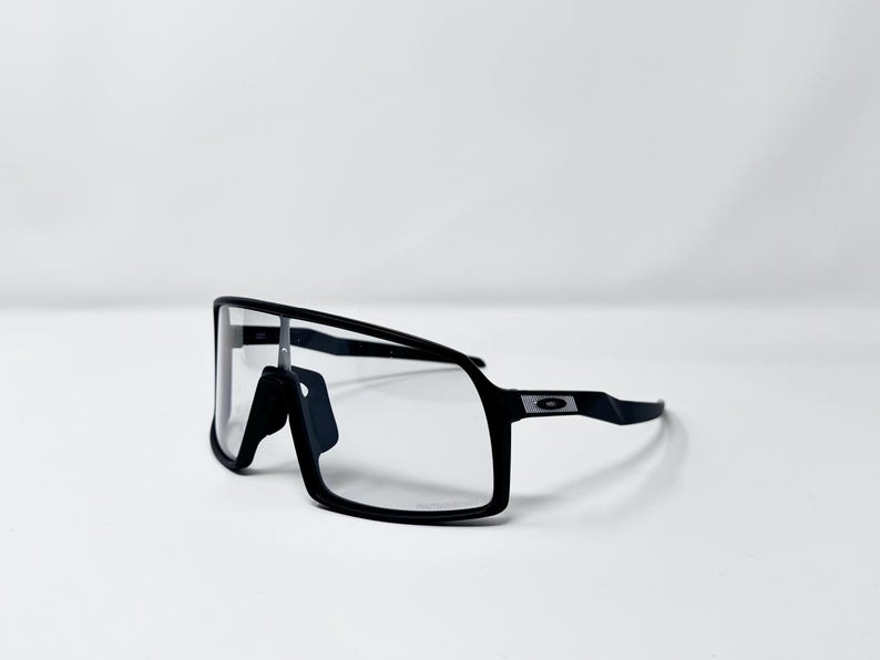 May include: Black sports sunglasses with a rectangular frame and clear lenses. The sunglasses have a sleek, modern design with a black frame and a small logo on the temple. The glasses are set against a white background.