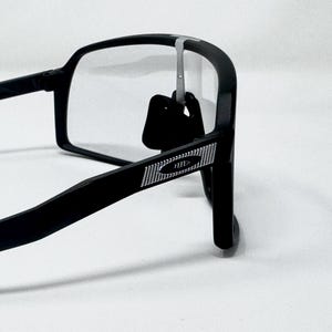 May include: Black rectangular eyeglasses with clear lenses. The frames have a matte finish and a silver accent on the temple. The glasses are shown against a white background.