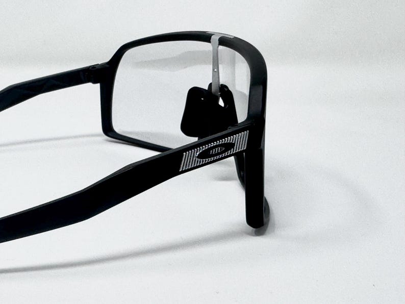 May include: Black rectangular eyeglasses with clear lenses. The frames have a matte finish and a silver accent on the temple. The glasses are shown against a white background.