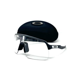 Op de afbeelding: Sportzonnebril met heldere glazen, zwart montuur en neuspads, voor een zwarte, ovale etui. De zonnebril heeft een modern design, geschikt voor buitenactiviteiten.