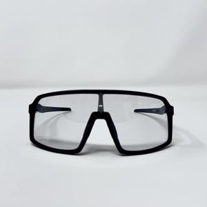 May include: Black sport sunglasses with a rectangular frame and clear lenses. The sunglasses have a sleek, modern design, suitable for outdoor activities. The frame is thick and wraps around the lenses.