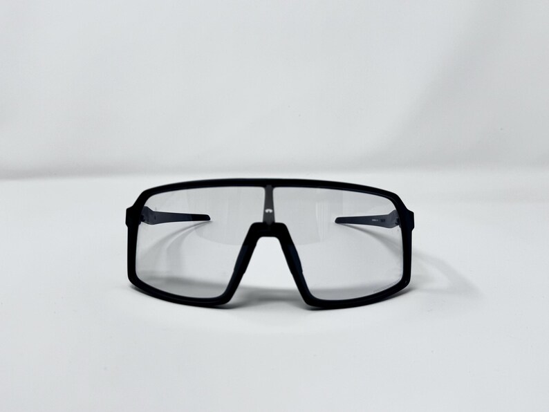 May include: Black sport sunglasses with a rectangular frame and clear lenses. The sunglasses have a sleek, modern design, suitable for outdoor activities. The frame is thick and wraps around the lenses.