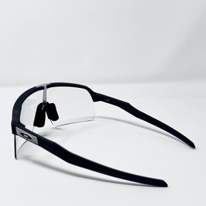 Op de afbeelding: Zwarte sportzonnebril met heldere glazen. De zonnebril heeft een strak, modern design met een matte afwerking. Het Oakley-logo is zichtbaar op het montuur. De glazen zijn aan de onderkant randloos.