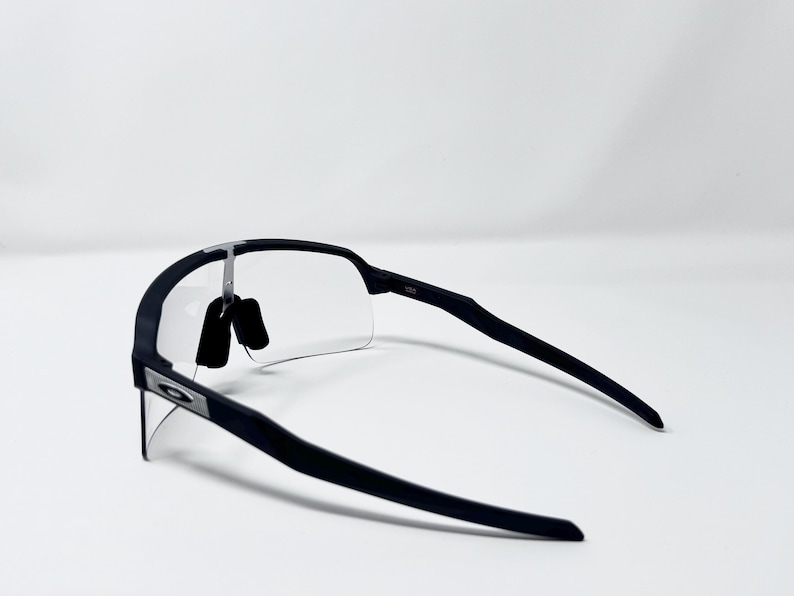 Op de afbeelding: Zwarte sportzonnebril met heldere glazen. De zonnebril heeft een strak, modern design met een matte afwerking. Het Oakley-logo is zichtbaar op het montuur. De glazen zijn aan de onderkant randloos.