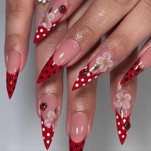 May include: Stiletto-shaped nails featuring a French manicure style with red tips adorned with white polka dots. The design includes three-dimensional white flower embellishments with gold centers and small red and black ladybug accents.