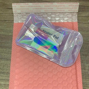 May include: A clear, iridescent pouch containing nail care items, including a tube of nail glue, a nail file, and an alcohol pad. The pouch is placed on a pink bubble mailer. The text "Alcohol Pad" is visible on the pad.