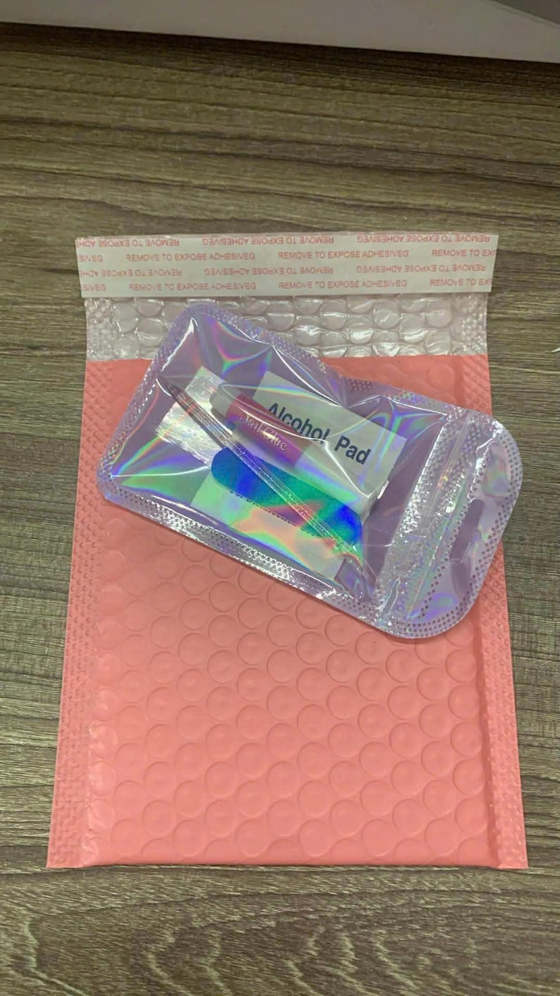 May include: A clear, iridescent pouch containing nail care items, including a tube of nail glue, a nail file, and an alcohol pad. The pouch is placed on a pink bubble mailer. The text "Alcohol Pad" is visible on the pad.