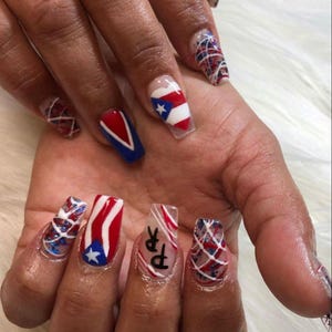 May include: A hand with acrylic nails featuring patriotic designs. The nails showcase the Puerto Rican flag, red and blue accents, and the letters "PR". Designs include stripes, stars, and geometric patterns.