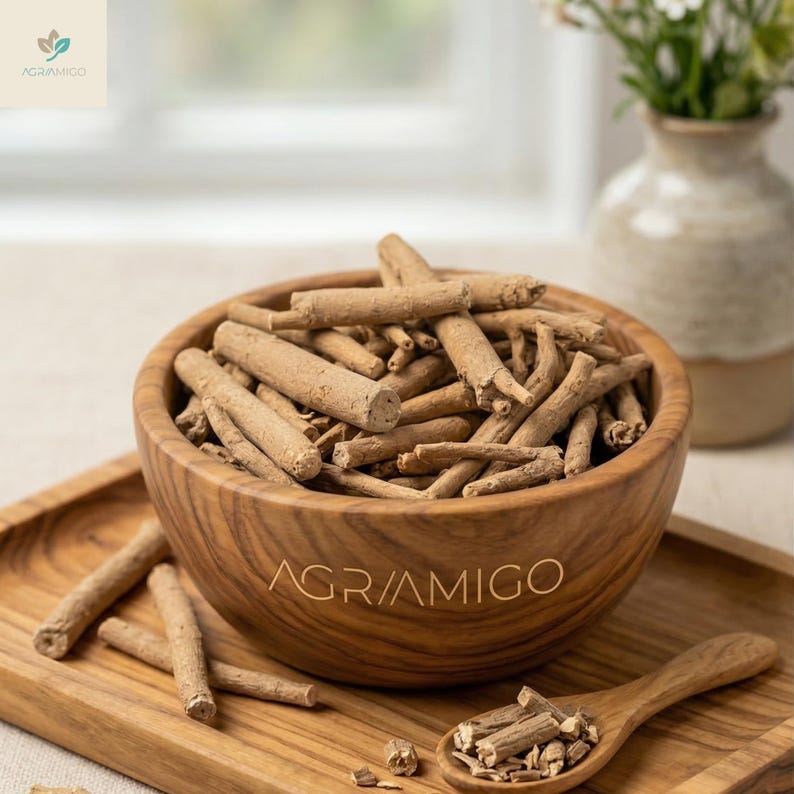May include: A wooden bowl filled with tan-colored ashwagandha root pieces, displayed on a wooden tray. A wooden spoon holds smaller pieces of the root. The bowl and tray have the text "AGRAMIGO" printed on them. A vase of flowers is in the background.