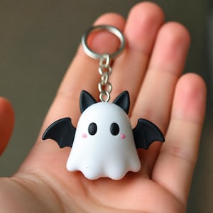 以下が含まれることがあります： 黒いコウモリの羽、黒い尖った耳、2つの真っ黒な楕円形の目、小さなピンクの丸い頬が特徴の白いお化けの形をしたキーホルダー。チャームは短い銀色の金属チェーンで銀色の金属製二重リングに取り付けられています。