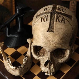 May include: A detailed, weathered skull with a cross carved into the forehead and the word "NIKA". The lower jaw is separate. A black object is in the background. The objects are on a checkered surface.