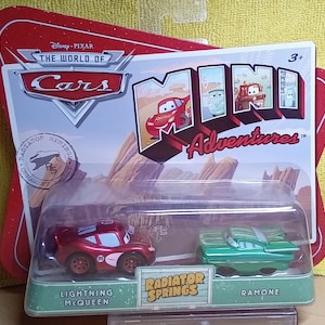 May include: A packaged set of two toy cars from the Disney Pixar Cars Mini Adventures series. The set includes a red Lightning McQueen car and a green Ramone car. The packaging features the Cars logo and the words "Radiator Springs".