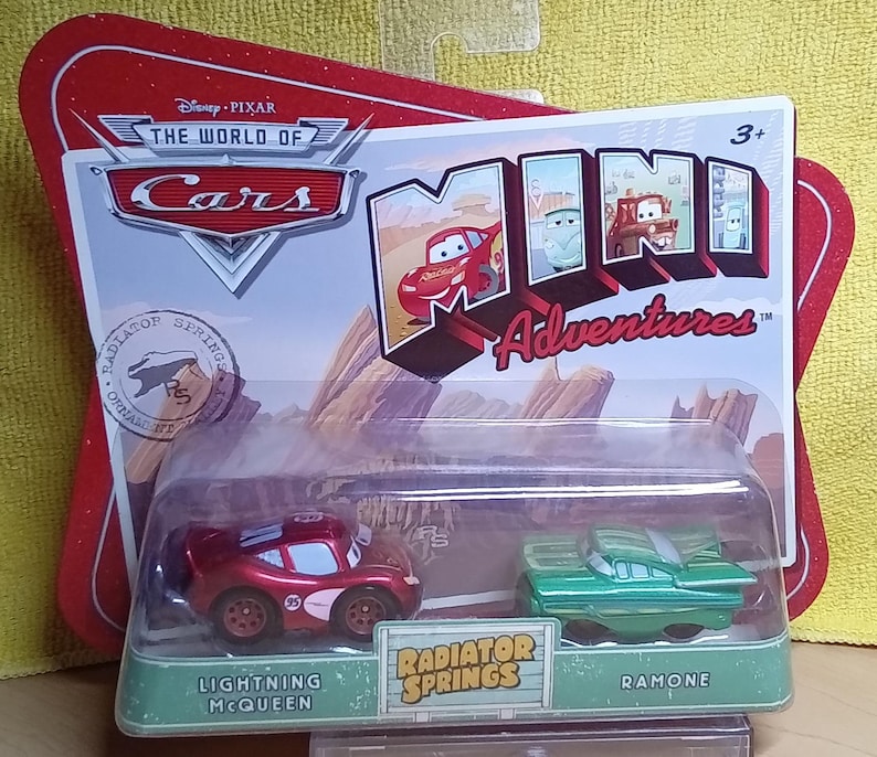 May include: A packaged set of two toy cars from the Disney Pixar Cars Mini Adventures series. The set includes a red Lightning McQueen car and a green Ramone car. The packaging features the Cars logo and the words "Radiator Springs".