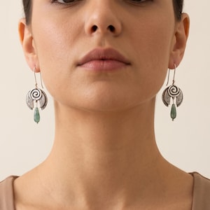 May include: A pair of silver dangle earrings featuring a circular spiral design with textured detailing. Each earring is finished with a small, teardrop-shaped green gemstone pendant suspended from the centre of the silver spiral component.