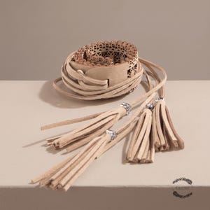 May include: A beige wrap belt featuring a laser-cut floral pattern along the main band. The belt includes long, thin leather-like ties that extend from the center, each finished with a dangling tassel accented by a small, square silver-toned bead.