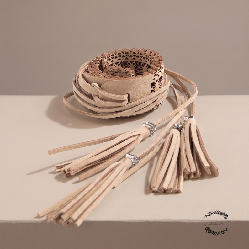 May include: A beige wrap belt featuring a laser-cut floral pattern along the main band. The belt includes long, thin leather-like ties that extend from the center, each finished with a dangling tassel accented by a small, square silver-toned bead.