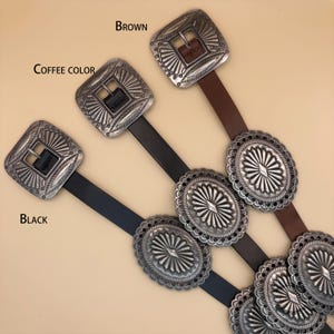 Puede incluir: Tres cinturones en negro, color caf&eacute; y marr&oacute;n, cada uno con una hebilla cuadrada plateada con grabado de rayos de sol y m&uacute;ltiples detalles ovalados de concho plateado a lo largo de la correa. Las etiquetas Black, Coffee color y Brown son visibles.