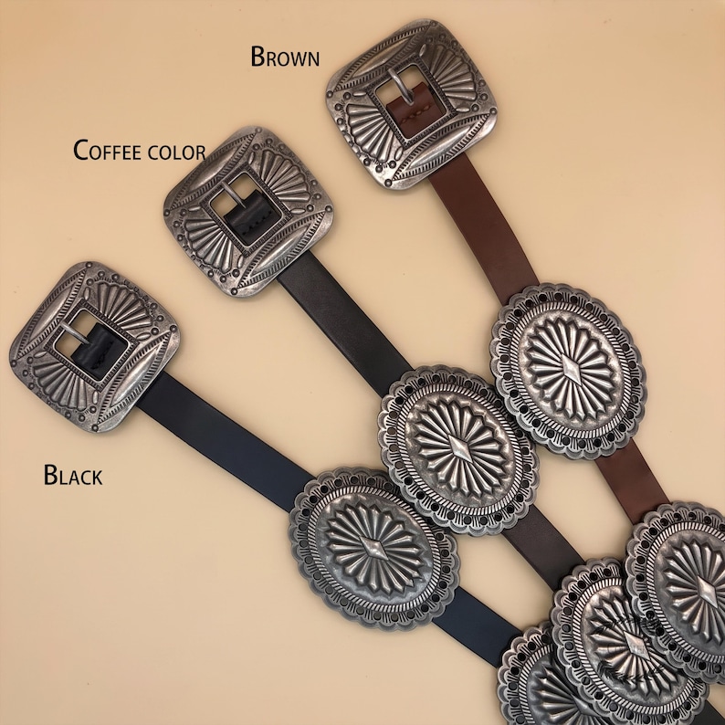 Puede incluir: Tres cinturones en negro, color caf&eacute; y marr&oacute;n, cada uno con una hebilla cuadrada plateada con grabado de rayos de sol y m&uacute;ltiples detalles ovalados de concho plateado a lo largo de la correa. Las etiquetas Black, Coffee color y Brown son visibles.