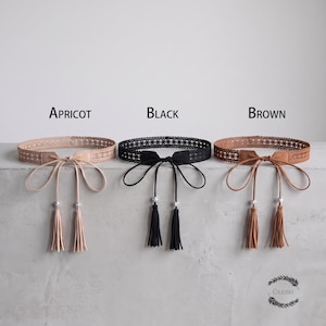 May include: Three belts in apricot, black, and brown, each featuring a wide, intricate lace-like band, a central tie closure with long, thin straps, and two dangling tassels accented with small, clear, reflective beads at the ends of each strap.