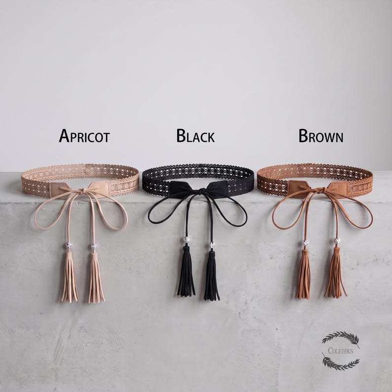 May include: Three belts in apricot, black, and brown, each featuring a wide, intricate lace-like band, a central tie closure with long, thin straps, and two dangling tassels accented with small, clear, reflective beads at the ends of each strap.