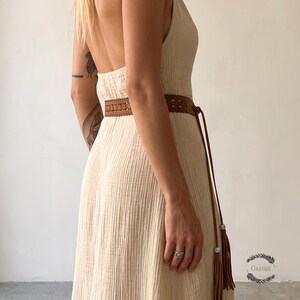 May include: A sleeveless, cream-colored, textured maxi dress with a low-cut back. A brown leather belt with intricate cut-out patterns and long, thin ties ending in metallic-accented tassels is wrapped around the waist of the dress.