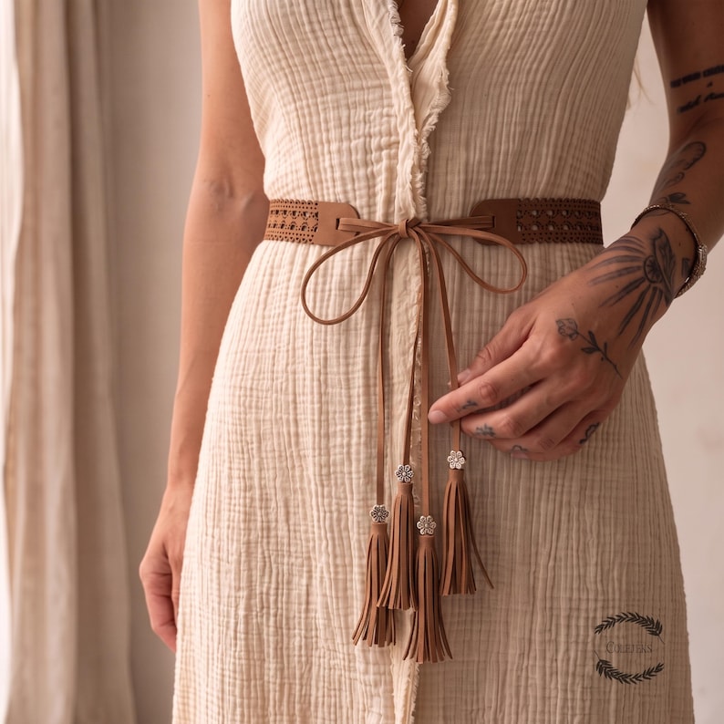 May include: A light beige sleeveless dress made of textured fabric featuring a raw edge down the center. A brown waist belt with a perforated pattern is tied in a bow at the front, with long dangling cords ending in silver flower charms and tassels.