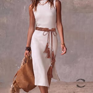 May include: A sleeveless white ribbed midi dress featuring a high neckline and a side slit. The dress is cinched at the waist with a brown perforated tie belt adorned with long dangling tassels and silver-toned beads. A large brown textured bag is held.