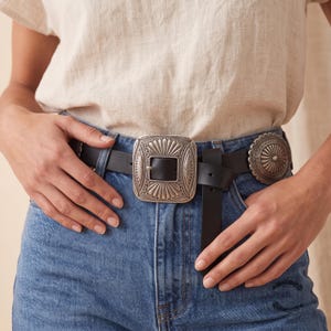 Puede incluir: Un cintur&oacute;n de cuero negro con una hebilla met&aacute;lica rectangular grande en tono plateado con patrones de rayos de sol grabados y un adorno circular a juego, usado con pantalones vaqueros azules y una blusa de lino de color claro.