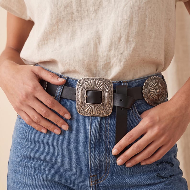 Puede incluir: Un cintur&oacute;n de cuero negro con una hebilla met&aacute;lica rectangular grande en tono plateado con patrones de rayos de sol grabados y un adorno circular a juego, usado con pantalones vaqueros azules y una blusa de lino de color claro.
