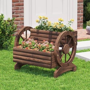 May include: A wooden planter shaped like a wagon with two large wheels. The planter is filled with colorful flowers and greenery. The wood has a dark brown stain, and the planter sits on a green lawn.