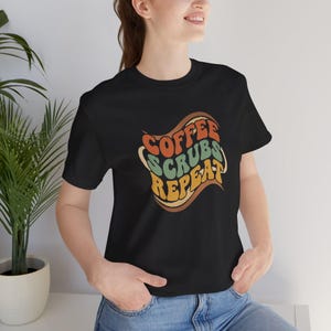 May include: Black t-shirt featuring the text "COFFEE SCRUBS REPEAT" in a retro design. The text is in orange, green, and yellow, with brown outlines. The shirt has a crew neck and short sleeves.
