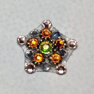 May include: A star-shaped embellishment with a clear, textured base. The star is adorned with various colorful rhinestones, including orange, green, blue, and pink, arranged in a decorative pattern. This item could be used for crafts or jewelry making.