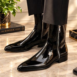 Könnte beinhalten: Ein Paar schwarze Lackleder-Stiefeletten mit Hochglanz-Finish, ausgestattet mit einer spitz zulaufenden Zehenpartie, einem niedrigen Blockabsatz und einem auffälligen silberfarbenen Metallreißverschluss an der Außenseite.