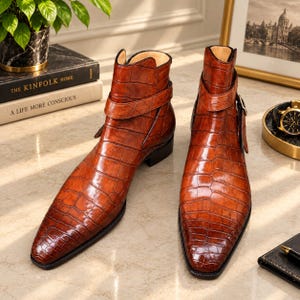 May include: A pair of brown ankle boots featuring a glossy crocodile skin texture, a pointed toe, and a decorative strap with a metal buckle around the ankle. The boots have a low heel and a smooth, polished finish, showcasing a rich tan colour.