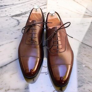 Könnte beinhalten: Ein Paar polierte dunkelbraune Oxford-Schuhe aus Leder mit Zehenkappe, dünnen braunen Schnürsenkeln und hellen Holzschuhspannern im Inneren. Die Schuhe zeichnen sich durch eine glatte, glänzende Oberfläche und eine strukturierte Form mit sichtbaren Nähten aus.
