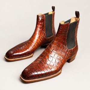Op de afbeelding: Een paar bruine Chelsea boots met een glanzende, in reliëf aangebrachte krokodillenleertextuur. Deze laarzen hebben een vierkante neus, een lage hak, zwarte elastische zijpanelen en een treklipje aan de bovenkant van de hielkraag.