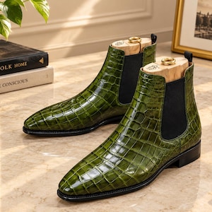 May include: A pair of olive green Chelsea boots featuring a glossy crocodile-embossed leather finish, black elastic side panels, and black pull tabs. Each boot contains a light-coloured wooden shoe tree with a gold-toned metal ring at the top.