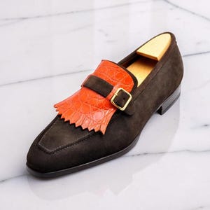 May include: A dark brown suede loafer featuring a bright orange crocodile-embossed leather kiltie fringe, a matching strap with a gold-toned metal buckle, and a light-colored wooden shoe tree inserted into the heel area of the footwear.