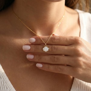 May include: A delicate gold necklace featuring a seashell pendant with a white center. The necklace is worn around the neck, showcasing the pendant's intricate design. The gold chain is thin and elegant, complementing the pendant's shape.