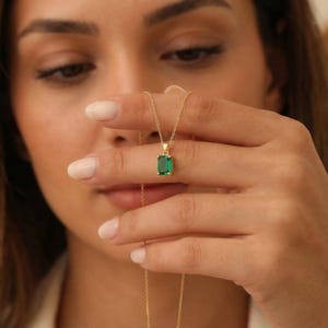 Può includere: Una collana color oro con un pendente rettangolare verde smeraldo. Il pendente è incastonato in una lunetta dorata e appeso a una delicata catena d'oro. La collana è mostrata su uno sfondo morbido.