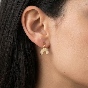 May include: Gold-colored rainbow earrings. The earrings feature a small, round stud that attaches to the ear, with a dangling rainbow pendant. The rainbow design has multiple curved lines, creating a textured effect. The earrings are worn on a person's ear.
